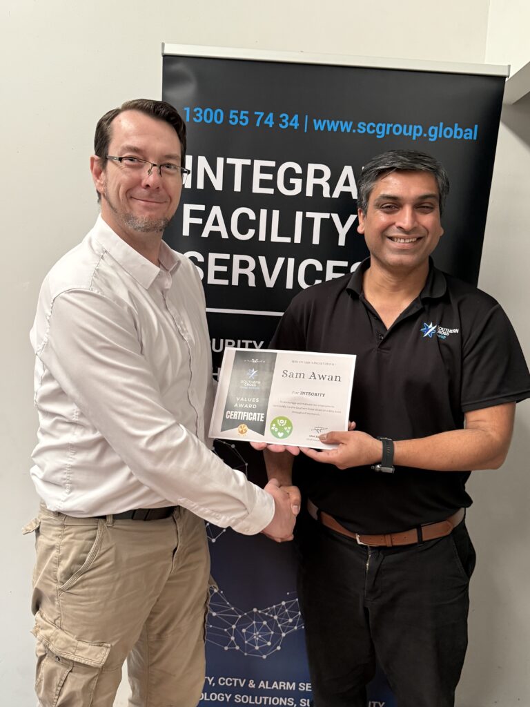Sam Awan stands holding a framed award certificate for Integrity while smiling. Stuart Moran, MBA, presents the award. Southern Cross Group branding is visible in the background.